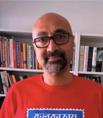 Imagem de um homem, o analista Bobby Gosh, vestido com uma t-shirt vermelha, com uma estante de livros atrás.