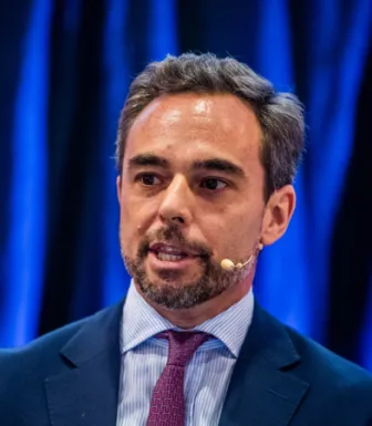 Retrato de um homem de fato com uma gravata roxa, a falar ao microfone.