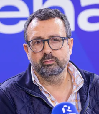 Retrato de um homem, sentado, a falar ao microfone, no estúdio da rádio Renascença.