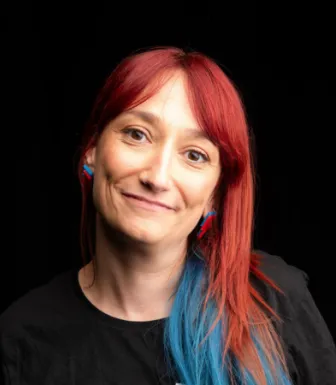 Retrato a cores da comunicadora Ana Markl, de cabelo vermelho e azul, vestida com uma t-shirt preta.