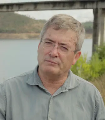 Retrato de um homem, de óculos, vestido com uma camisa. Uma barragem de fundo.