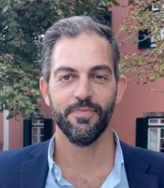 Imagem de um homem, com barba. Camisa e casaco azuis.