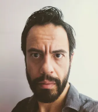 Retrato a cores de um homem, de barba e cabelo escuro, vestido com uma camisa cinzenta, sobre uma parede branca.