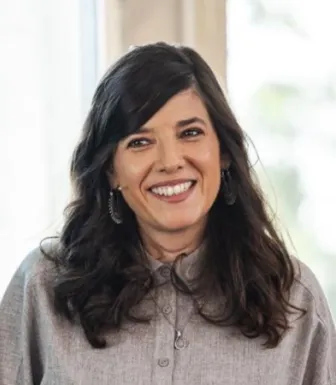 Retrato a cores de uma mulher sorridente, de cabelo comprido e escuro, vestida com uma camisa cinzenta. Fundo claro desfocado.