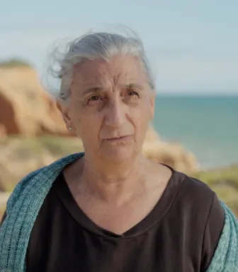 Retrato de uma senhora de cabelo branco, vestida com uma t-shirt preta. Ao fundo, o mar.