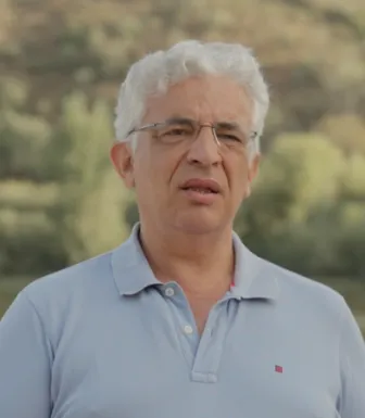 Retrato a cores de um homem de cabelo branco, com óculo, e vestido com um polo azul claro. Um rio e vegetação cobrem o fundo.