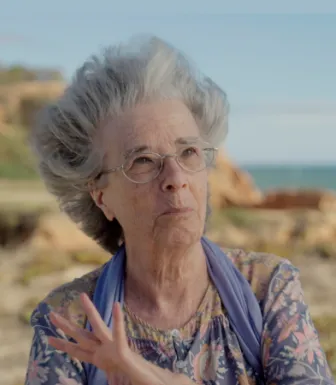Retrato a cores de uma senhora idosa, que usa óculos e um lenço ao pescoço. Uma praia cobre o fundo.