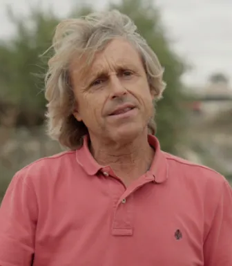 Retrato a cores de um homem com cabelo claro e pelos ombros, vestido com um polo rosa. 
