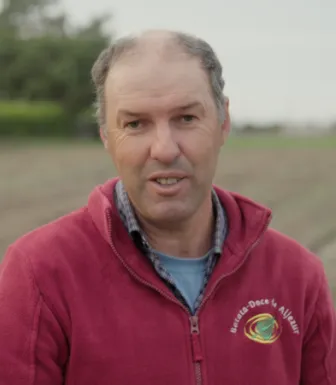 Retrato a cores de um homem, vestido com um casaco vermelho onde se lê "Batata-doce de Aljezur». Um campo agrícola cobre o fundo.