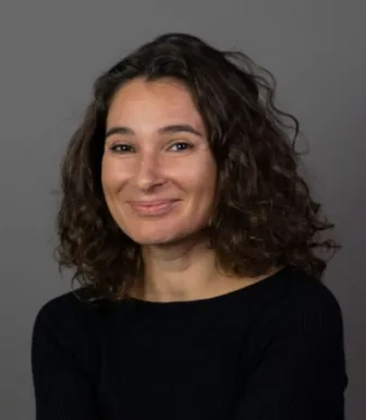 Retrato a cores de uma mulher, sorridente, de cabelo ondulado e escuro, vestida com uma camisola preta. Fundo cinzento escuro.