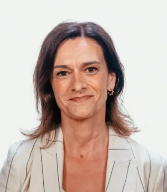 Retrato de uma mulher, sorridente, de cabelo castanho, vestida com um top branco e um blazer bege com riscas pretas. DR