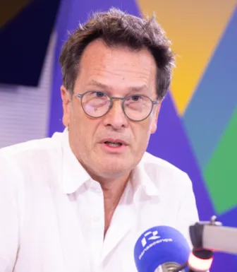 Um homem de óculos, num estúdio de rádio, a falar para um microfone. Fundo com várias cores.