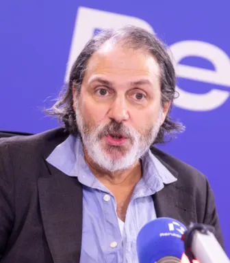 Retrato a cores de um homem com barba e cabelo grisalhos, vestido com uma camisa azul e um blazer preto, a falar para um microfone.