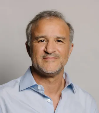 Retrato a cores de um homem, de cabelo grisalho, expressão sorridente. Veste uma camisa azul clara. DR