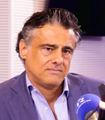 Um homem, vestido com um fato azul escuro e uma camisa azul clara, num estúdio de rádio, com um microfone à frente. Créditos: Fábio Oliveira.