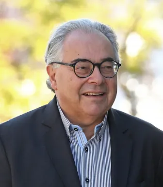 Retrato a cores de um homem de cabelo grisalho e óculos. Veste um blazer preto e uma camisa às riscas.