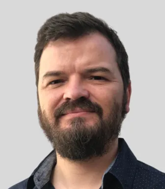Retrato a cores de homem sorridente; tem barba e cabelo castanhos e veste uma camisa escura. Fundo cinzento.
