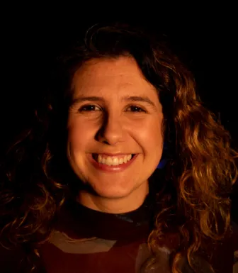 Retrato a cores de uma mulher, sorridente, de cabelo castanho encaracolado. Fundo preto.