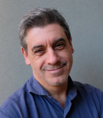 Retrato a cores de um homem, sorridente, de barba e cabelo grisalhos, vestido com uma camisa azul. Fundo cinzento.