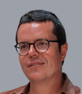 Retrato a cores de um homem, de óculos e cabelo escuro e curto, a sorrir. Fundo cinzento.