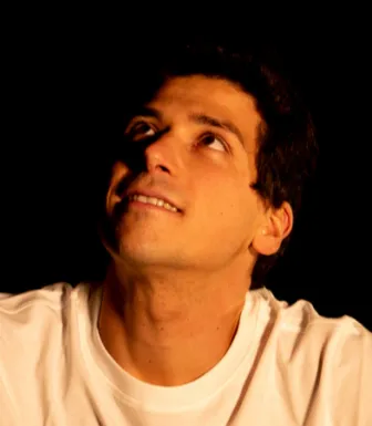 Retrato a cores de um homem sorridente a olhar para cima; veste uma t-shirt branca. Fundo preto.