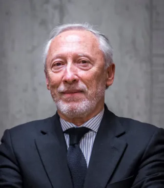 Retrato a cores de um homem com cabelo e barba grisalhos, vestido com um fato e gravata pretos. Fundo em pedra.
