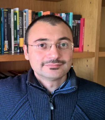 Retrato a cores de um homem que usa óculos e veste um camisola azul. Uma estante com livros ao fundo.