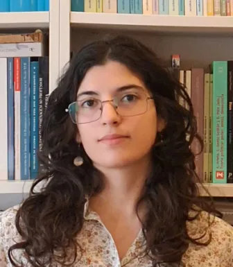 Retrato a cores de uma rapariga jovem de óculos e cabelo escuro ondulado. Uma estante de livros cobre o fundo.