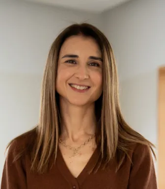 Retrato a cores de uma mulher sorridente, que veste um casaco castanho e usa um colar ao pescoço.