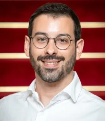 Retrato a cores de um homem com barba e de óculos, a sorrir, vestido com uma camisa branca. Fundo vermelho com riscas brancas. 
