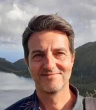 Retrato a cores de um homem a sorrir, com um lago e montanhas ao fundo.