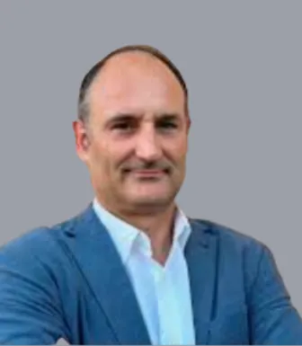 Retrato a cores de um homem vestido com uma camisa branca e um blazer azul. Fundo cinzento.D/R