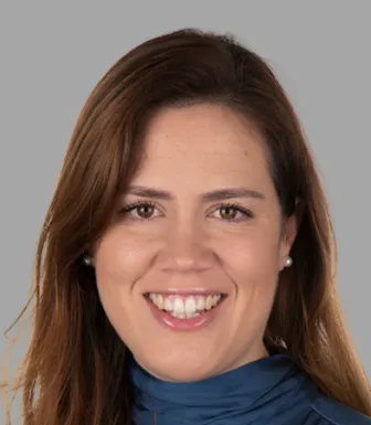 Retrato a cores de uma mulher sorridente, de cabelo castanho e comprido, e veste uma camisola azul. Fundo cinzento.