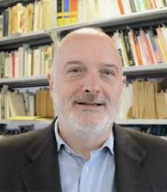 Retrato a cores de um homem com barba branca. Uma estante de livros cobre o fundo. D/R