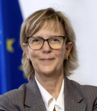 Retrato a cores de uma mulher com expressão sorridente. Tem cabelo claro e usa óculos. Bandeira da UE ao fundo.