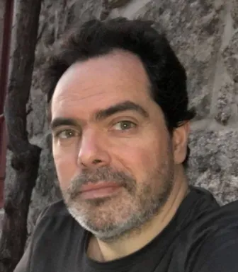 Retrato a cores de um homem com barba grisalha e cabelo escuro. Veste uma t-shirt preta. Fundo de uma parede em pedra.