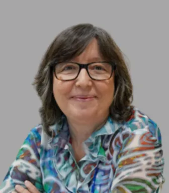 Retrato de uma mulher sorridente, com óculos e cabelo escuro. Está de braços cruzados e usa uma camisa colorida.