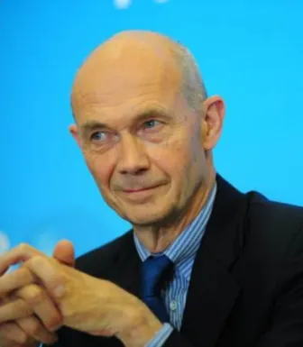 Retrato a cores de um homem, Pascal Lamy, com as mãos cruzadas. Veste um fato preto e uma camisa azul. Fundo azul claro.
