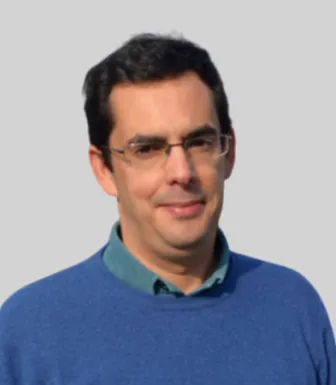Retrato a cores de um homem a sorrir, que usa óculos e tem cabelo escuro. Veste uma camisola azul. Fundo cinzento.