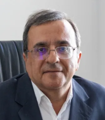 Retrato a cores de um homem com óculos, casaco preto e camisa branca, sentado numa cadeira de escritório. D/R