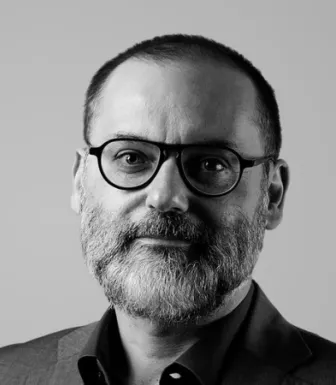 Retrato a preto e brancdo de um homem com barba, cabelo curto. Usa fato e óculos. 