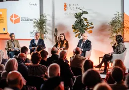 Uma plateia sentada, de costas, a olhar para um palco onde estão quatro oradores, entre eles Pedro Passos Coelho e Elvira Fortunato.