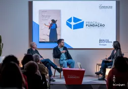 Uma plateia, de costas, a olhar para um palco, onde estão dois homens sentados a falar.