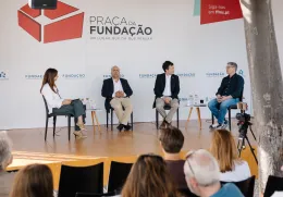 Quatro pessoas em palco, sentadas em cadeiras pretas, a falar com plateia a assistir. Logo e texto "Praça da Fundação".