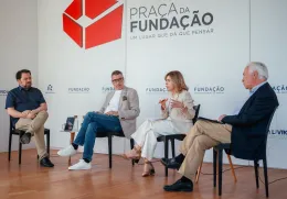 Quatro pessoas sentadas em cadeiras pretas, a debater entre si. Ao fundo, o texto "Praça da Fundação"