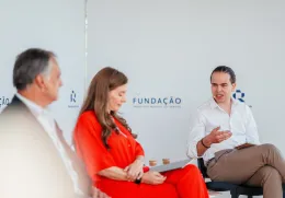 Três pessoas sentadas num palco. Dois homens de fato, uma senhora com um vestido vermelho. Ao fundo, o logotipo da Fundação.