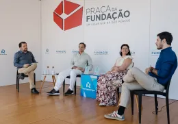 Quatro pessoas sentadas num palco, em cadeiras pretas, a apresentar um livro que está pousado em cima de uma mesa. Fundo com logotipos da Fundação.