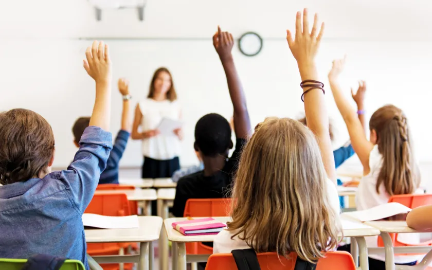 Imagem de alunos, sentados numa sala de aula, com o braço no ar para responderem à professora.