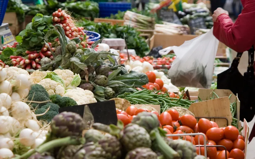 Imagem de mulher a comprar vegetais numa praça. Crédito: Canva