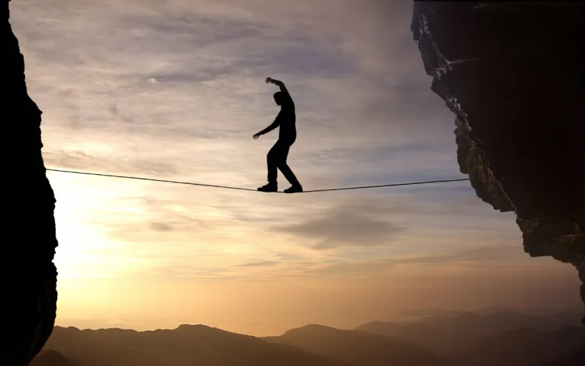 Imagem de um homem a caminhar numa arame suspenso entre duas rochas. Crédito: Canva